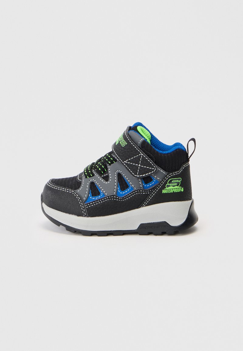 Schwarzer und blauer wasserdichter Kindersneaker mit grünen Akzenten, Klettverschluss und dicker weißer Sohle auf einfarbigem Hintergrund.