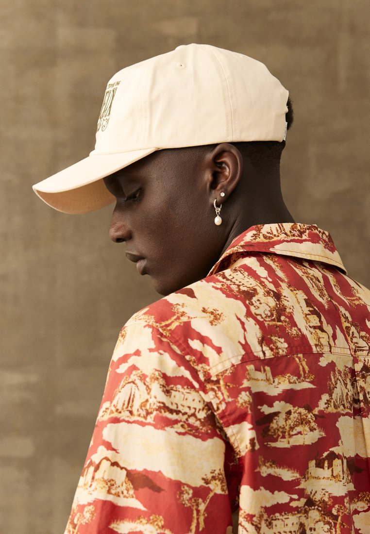 Beige Baseball-Cap und rotes, gemustertes Hemd mit Landschaftsprint, aus glattem Stoff mit kontrastierenden erdigen Farbtönen.