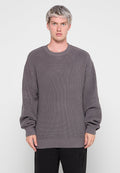 Grauer Strickpullover mit gerippter Textur, Rundhalsausschnitt und übergroßen Ärmeln. Mit einem Logo am linken Ärmel. Kombiniert mit dunklen Hosen.