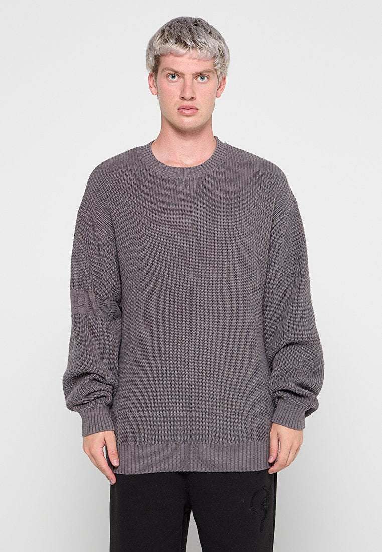 Grauer Strickpullover mit gerippter Textur, Rundhalsausschnitt und übergroßen Ärmeln. Mit einem Logo am linken Ärmel. Kombiniert mit dunklen Hosen.