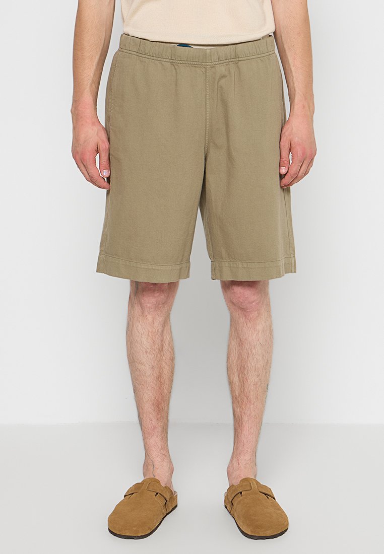 Beige Baumwollshorts mit elastischem Taillenbund, knielang, mit seitlichen Taschen und einem sauberen Saum. Kombiniert mit braunen Schlupfschuhen.