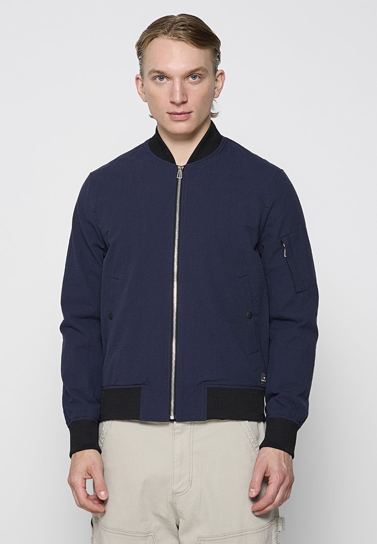 Marineblauer Bomberjacke mit Reißverschluss vorne, gerippten Bündchen und Saum. Verfügt über Seitentaschen und eine mit Reißverschluss versehenen Armtasche für zusätzliche Funktionalität.