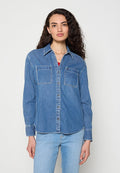 Frau mit langen lockigen Haaren, die ein blaues Jeanshemd mit Knöpfen und hellblaue Jeans vor einem einfarbigen weißen Hintergrund trägt.