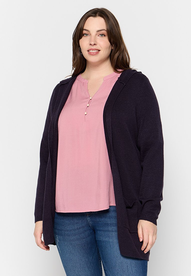 Marinefarbener Cardigan mit Kapuze, offene Front, gerippte Bündchen, getragen über einem pinken Oberteil mit Knopfleiste, kombiniert mit blauen Jeans. Glatte Stofftextur.