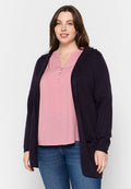 Marinefarbener Cardigan mit Kapuze, offene Front, gerippte Bündchen, getragen über einem pinken Oberteil mit Knopfleiste, kombiniert mit blauen Jeans. Glatte Stofftextur.