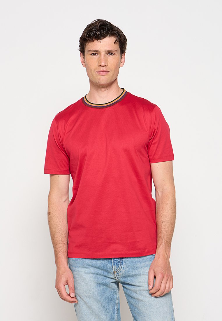 Mann mit kurzen braunen Haaren steht frontal, trägt ein rotes T-Shirt und hellblaue Jeans vor weißem Hintergrund.