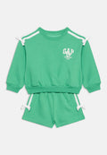 Grünes zweiteiliges Kinderoutfit mit langärmeligem Oberteil und Shorts, weißen Schulterstreifen, Schleifen und dem Schriftzug "GAP Friendship Club" mit Blumen.