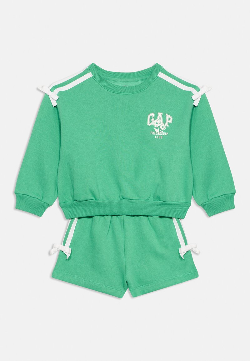 Grünes zweiteiliges Kinderoutfit mit langärmeligem Oberteil und Shorts, weißen Schulterstreifen, Schleifen und dem Schriftzug "GAP Friendship Club" mit Blumen.