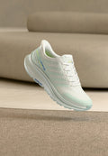 Weißer und hellgrüner Laufschuh mit Schnürsenkeln, der mit "Skechers GO RUN" gebrandet ist, schwebt über einem beigen Teppich und einer passenden Sofahintergrund.