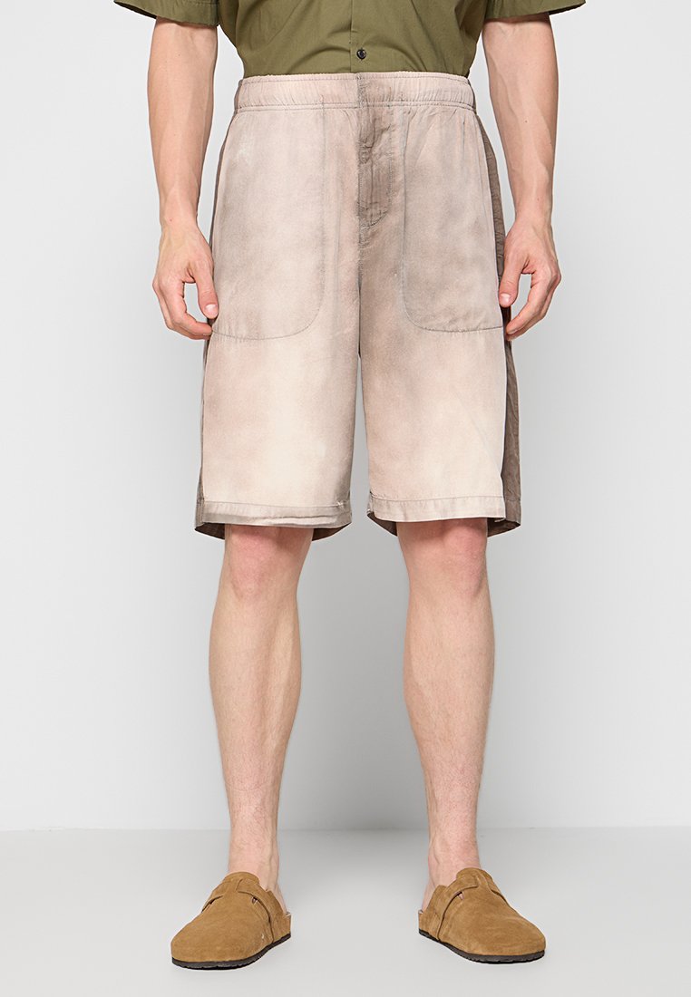Beige Shorts mit einem verwitterten, strukturierten Finish, ausgestattet mit zwei großen Seitentaschen und einem elastischen Bund. Getragen mit braunen Slippers.
