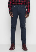 Mann trägt dunkelblaue Slim-Fit-Jeans, rot-schwarzes kariertes Hemd und glänzende bordeauxrote Sneaker, steht vor einem einfarbigen weißen Hintergrund.