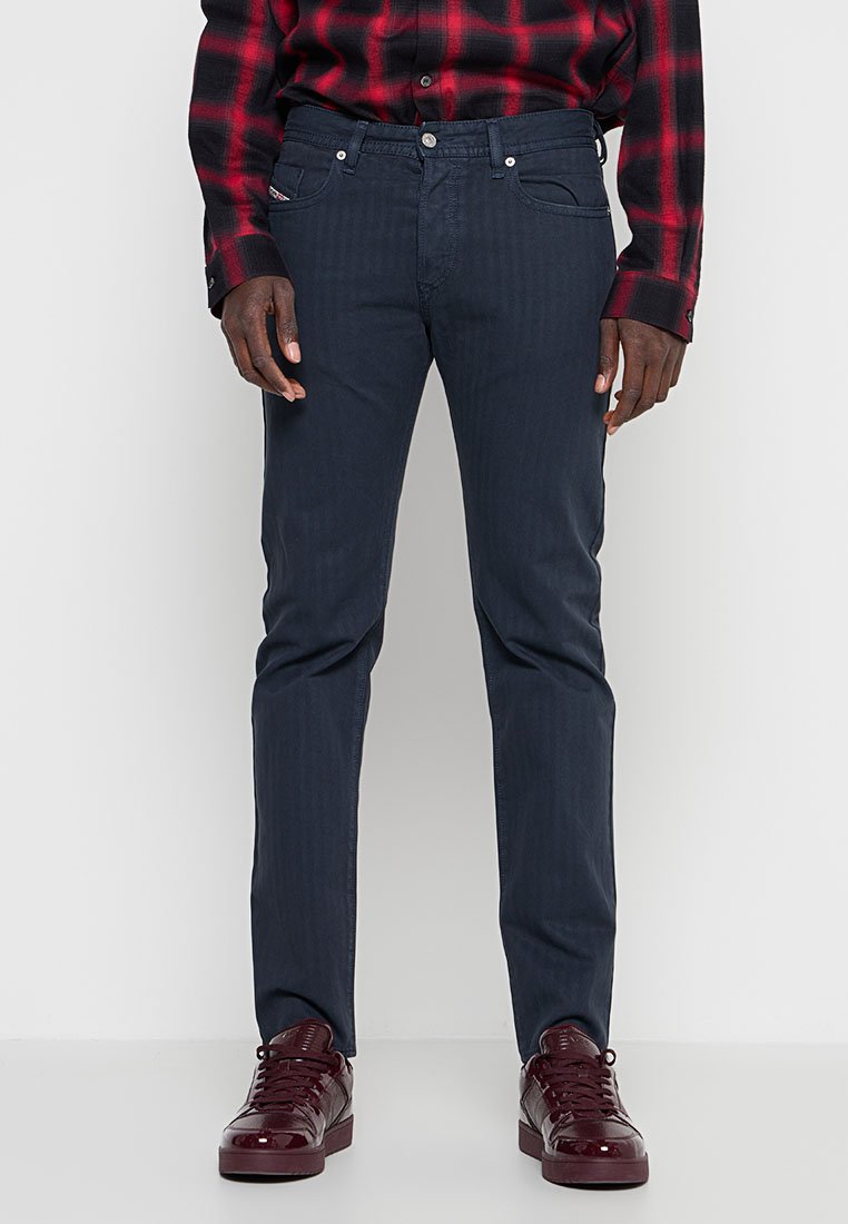 Mann trägt dunkelblaue Slim-Fit-Jeans, rot-schwarzes kariertes Hemd und glänzende bordeauxrote Sneaker, steht vor einem einfarbigen weißen Hintergrund.