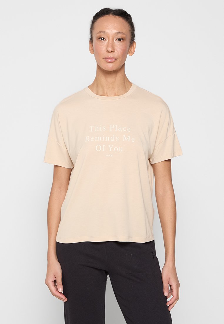Beige T-Shirt mit Kurzarm und Rundhalsausschnitt, auf dem weißen Text steht: „Dieser Ort erinnert mich an dich.“ Weicher Stoff und lockere Passform.