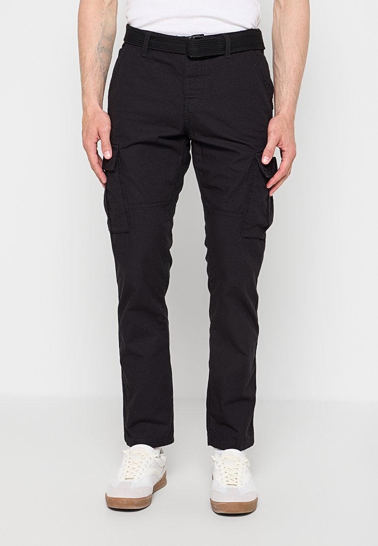Mann trägt schwarze Cargohosen mit Seitentaschen, einen schwarzen Gürtel, weiße Sneaker und ein weißes Hemd, steht vor einem schlichten hellen Hintergrund.