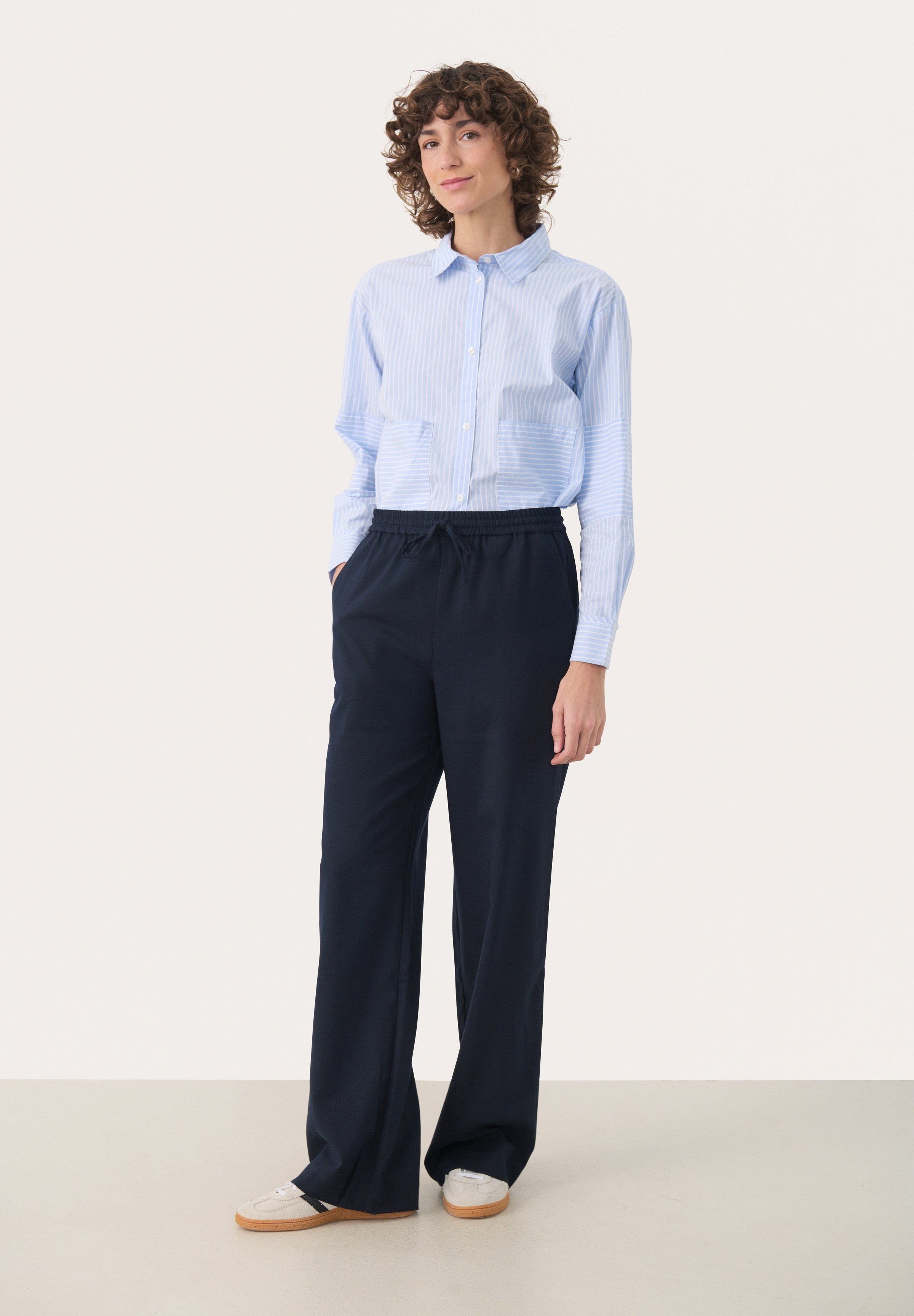 Blau-weiß gestreiftes Hemd mit Knöpfen und zwei aufgesetzten Taschen vorne, kombiniert mit navyblauen elastischen Hosen und lässigen weißen Sneakers.