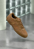 Brauner Wildleder-Sneaker mit "HUGO"-Logo an der Seite, detaillierter Naht und Schnürsenkeln, schwebend über einem polierten Betonboden vor einer Glasblockwand.