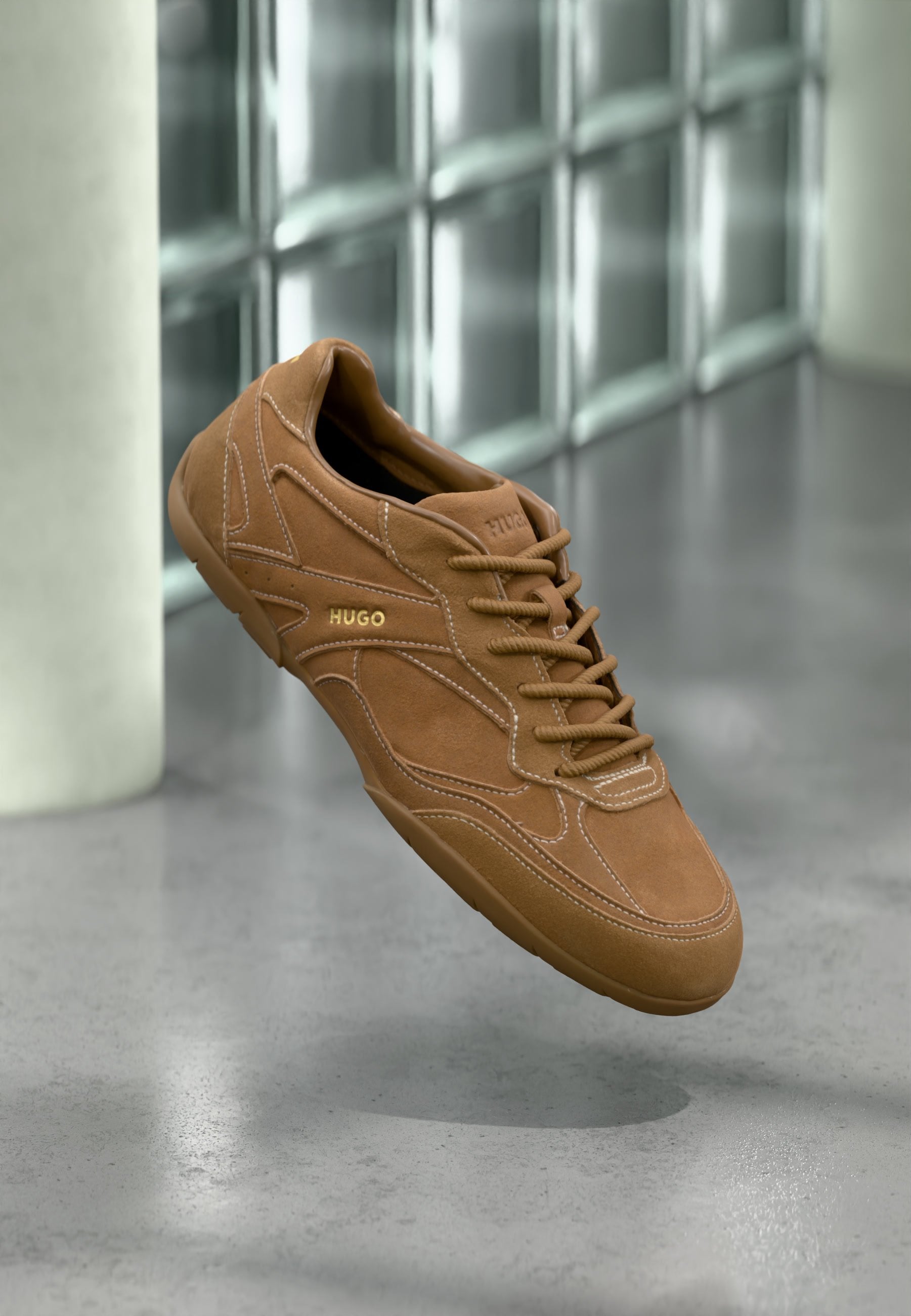Brauner Wildleder-Sneaker mit "HUGO"-Logo an der Seite, detaillierter Naht und Schnürsenkeln, schwebend über einem polierten Betonboden vor einer Glasblockwand.