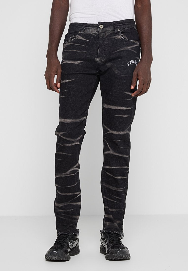 Mann trägt schwarze Acid-Wash-Jeans mit dem Text "PEGAD" auf dem Oberschenkel und schwarze Sneakers, steht vor einem schlichten weißen Hintergrund.