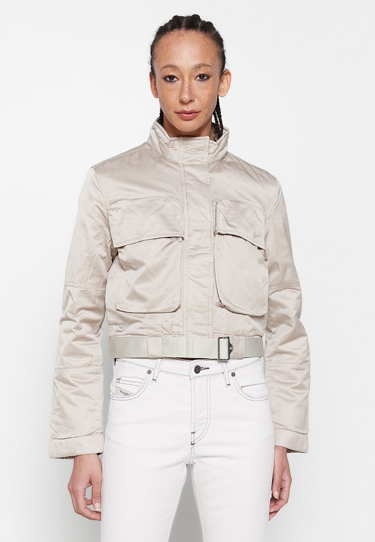 Beige cropped Jacke mit hohem Kragen, zwei frontalen Taschen und passendem Gürtel-Detail. Glattes, leichtes Material mit subtiler Schattierung.