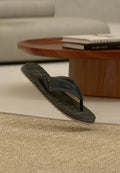 Ein einzelner schwarz-grauer Flip-Flop mit strukturiertem Absatz, der über einem gewebten beigen Teppich in einem modernen Wohnzimmer schwebt.