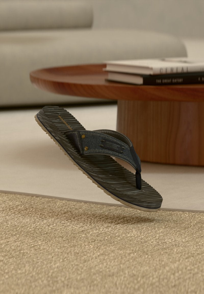 Ein einzelner schwarz-grauer Flip-Flop mit strukturiertem Absatz, der über einem gewebten beigen Teppich in einem modernen Wohnzimmer schwebt.