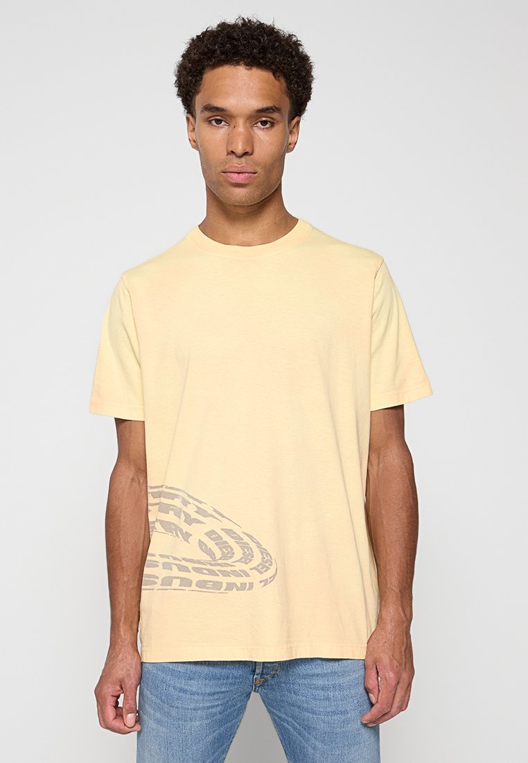 Mann mit lockigem Haar, der ein blassgelbes T-Shirt mit grauem Textdesign unten links trägt, kombiniert mit hellblauen Jeans.