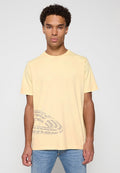 Mann mit lockigem Haar, der ein blassgelbes T-Shirt mit grauem Textdesign unten links trägt, kombiniert mit hellblauen Jeans.