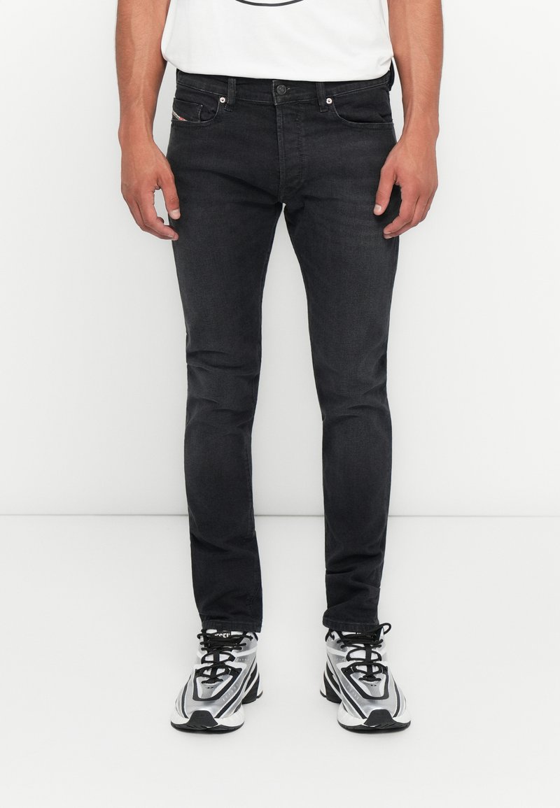 Mann in slim-fit schwarzen Jeans und schwarz-weißen Sportschuhen, der vor einem weißen Hintergrund steht.