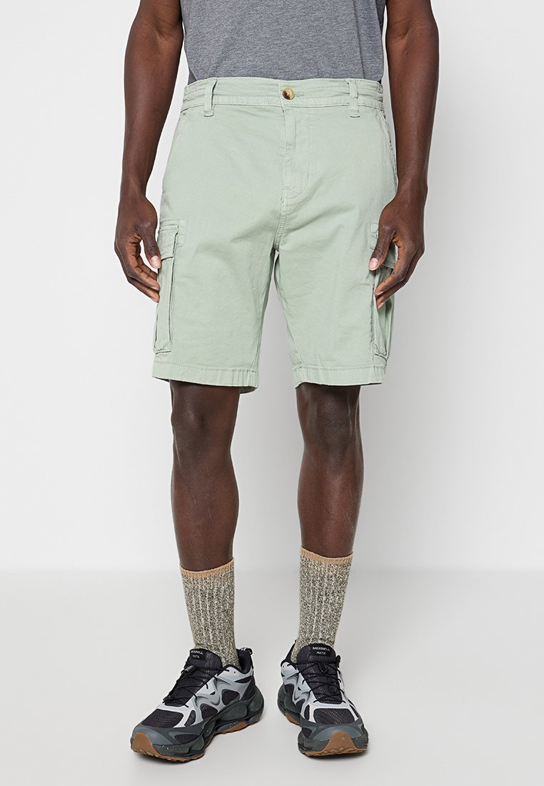 Mann trägt hellgrüne Cargo-Shorts, ein graues T-Shirt, grau-beige Socken und schwarze, graue sowie weiße Sneaker und steht vor einem schlichten Hintergrund.