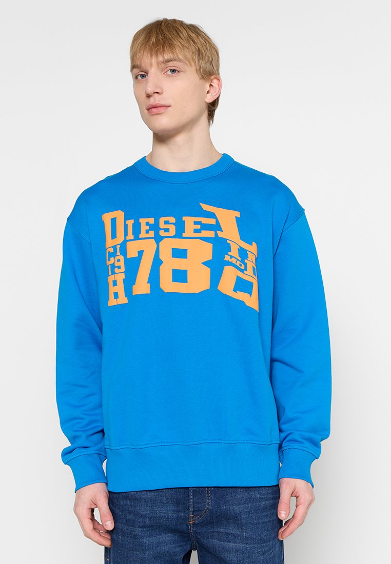 Junger Mann steht in einem leuchtend blauen Sweatshirt mit großen orangen "DIESEL 78"-Aufdruck und dunkelblauen Jeans vor einem weißen Hintergrund.