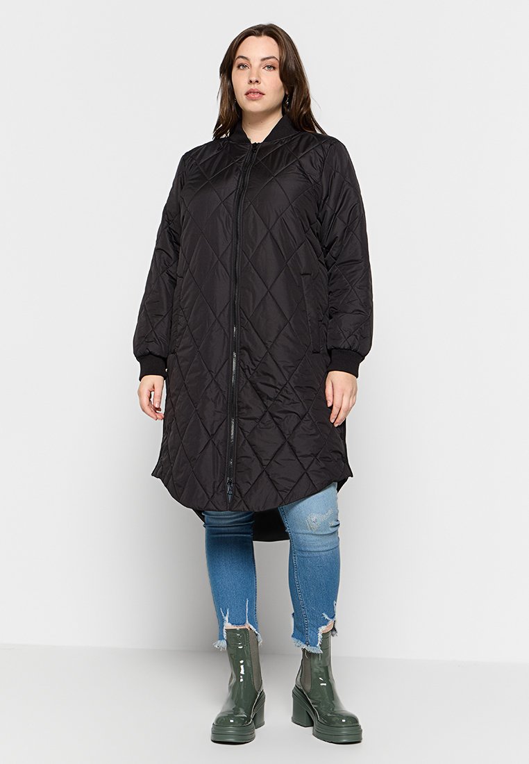 Schwarzer gesteppter langer Mantel mit Reißverschluss, gerippten Bündchen und abgerundetem Saum. Getragen über verwaschenen blauen Jeans und grünen Stiefeletten.