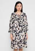 Frau, die ein lockeres, langärmeliges Kleid in knielanger Länge mit einem abstrakten Blumenmuster in Beige, Schwarz und Hellblau vor einem einfarbigen Hintergrund trägt.