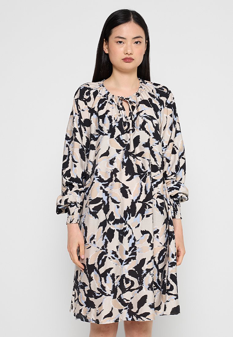 Frau, die ein lockeres, langärmeliges Kleid in knielanger Länge mit einem abstrakten Blumenmuster in Beige, Schwarz und Hellblau vor einem einfarbigen Hintergrund trägt.
