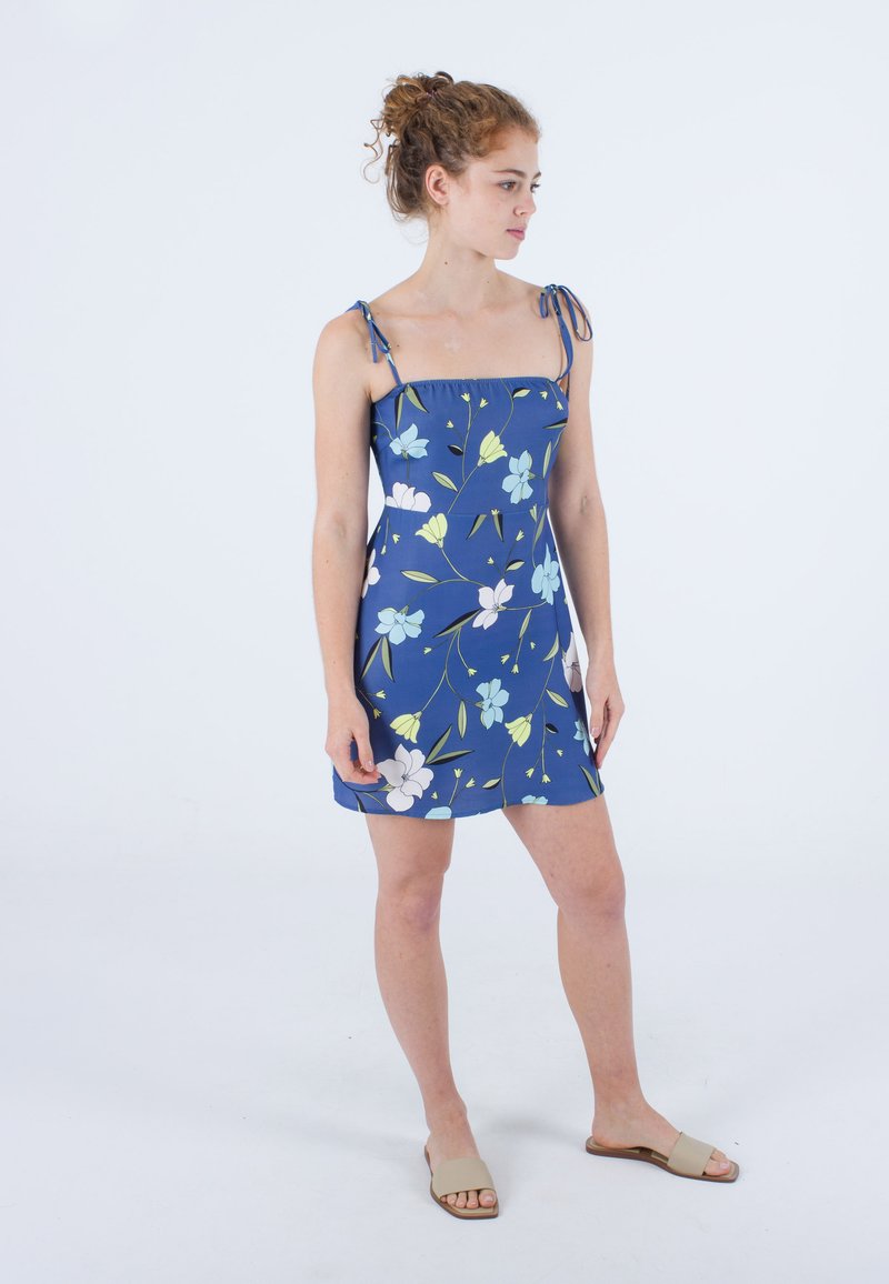 Kurzes blaues Kleid mit floralem Muster in Weiß und Grün, verstellbaren Bindeträgern und passgenauem Oberteil, sitzt oberhalb der Knie.