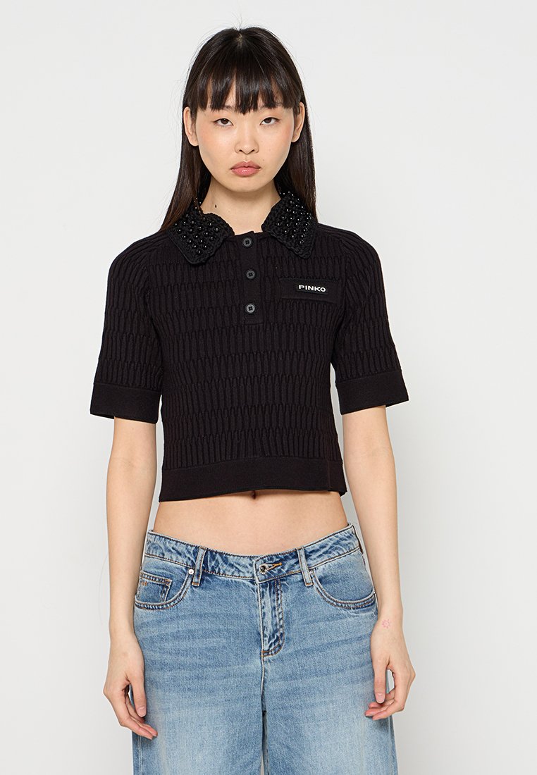 Junge Frau in einem schwarzen, strukturierten, kurzärmeligem Crop-Top mit beaded Kragen und blauen, hoch taillierten Jeans, die vor einem schlichten Hintergrund steht.