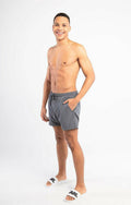Graue Schwimmshorts mit elastischem Bund und Seitentaschen, kombiniert mit weißen Slides mit schwarzem Text. Das Modell steht vor einem neutralen Hintergrund.