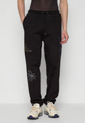 Schwarze Jogginghosen mit elastischem Bund, Kordelzug und gestickten Designs von Pflanzen in Weiß und Gold an den Seiten. Getragen mit beigen Sneakers.