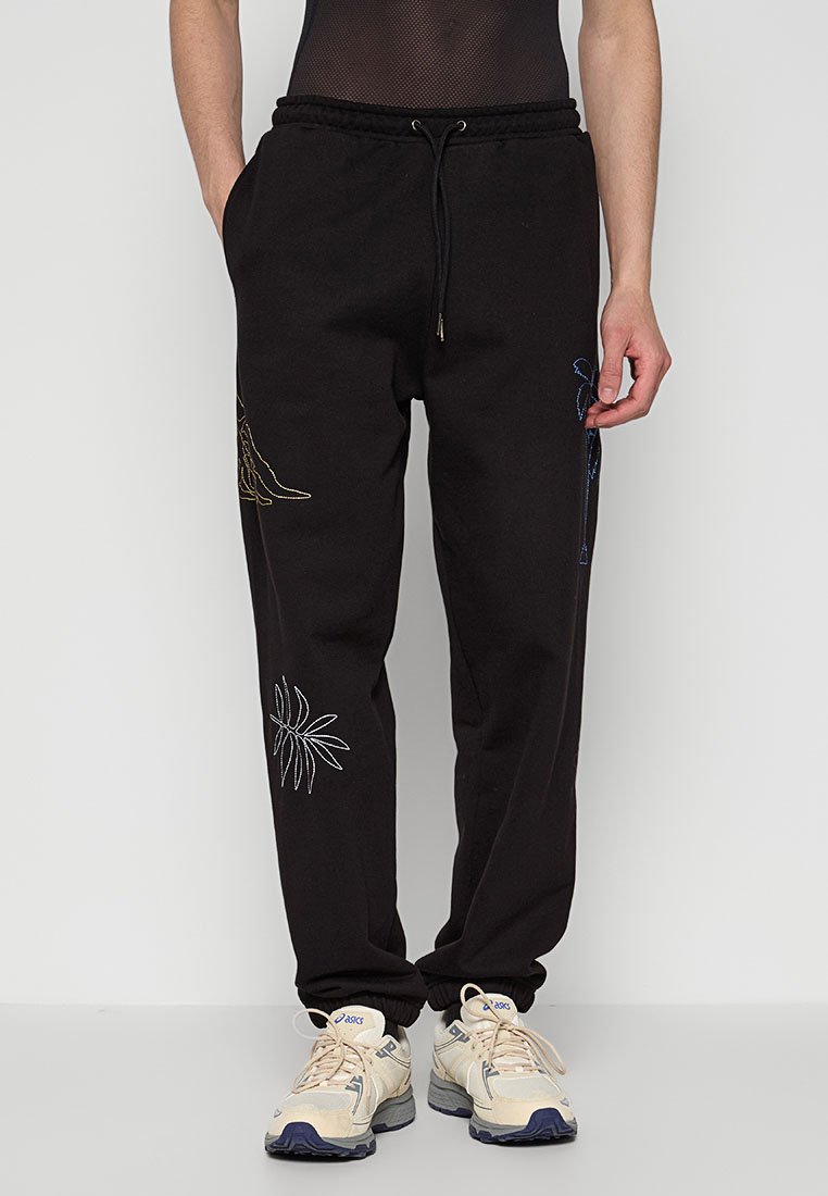 Schwarze Jogginghosen mit elastischem Bund, Kordelzug und gestickten Designs von Pflanzen in Weiß und Gold an den Seiten. Getragen mit beigen Sneakers.