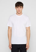 Junger Mann mit kurzen braunen Haaren, der ein einfaches weißes T-Shirt mit kurzen Ärmeln und schwarze Hose trägt, steht vor einem hellgrauen Hintergrund.