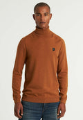 Ein rostfarbener Rollkragenpullover aus weichem Stoff, mit schmaler Passform und gerippten Bündchen, versehen mit einem kleinen schwarzen Logo auf der Brust. Kombiniert mit blauen Jeans.