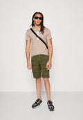 Junger Mann mit langen Dreadlocks, der schwarze Sonnenbrillen, beige T-Shirt, grüne Cargoshorts, Umhängetasche und mehrfarbige Sneakers trägt und vor einer weißen Wand steht.