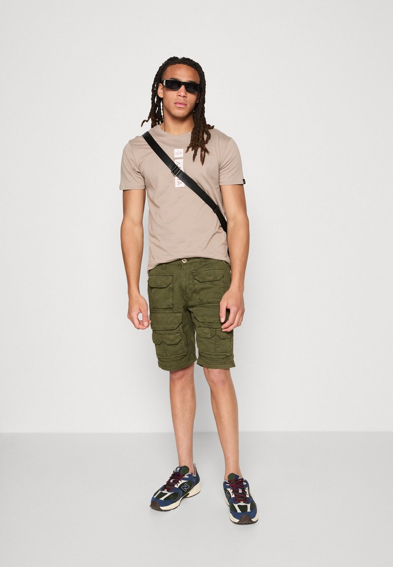 Junger Mann mit langen Dreadlocks, der schwarze Sonnenbrillen, beige T-Shirt, grüne Cargoshorts, Umhängetasche und mehrfarbige Sneakers trägt und vor einer weißen Wand steht.