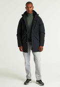Dunkelblauer Parka mit Reißverschluss und Druckknopfverschluss, Kapuze und mehreren Taschen; kombiniert mit einem grünen Rollkragenpullover und hellen Jeans.