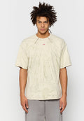 Junger Mann mit dunklem, lockigem Haar, der ein beiges, mit Batikmuster versehenes, übergroßes T-Shirt und graue Hosen trägt und vor einem schlichten weißen Hintergrund steht.