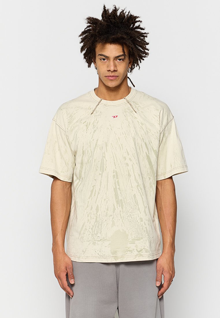 Junger Mann mit dunklem, lockigem Haar, der ein beiges, mit Batikmuster versehenes, übergroßes T-Shirt und graue Hosen trägt und vor einem schlichten weißen Hintergrund steht.