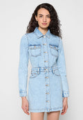 Frau mit lockigem Haar, die ein hellblaues Jeanskleid mit langen Ärmeln und Brusttaschen trägt und vor einem schlichten Hintergrund steht.