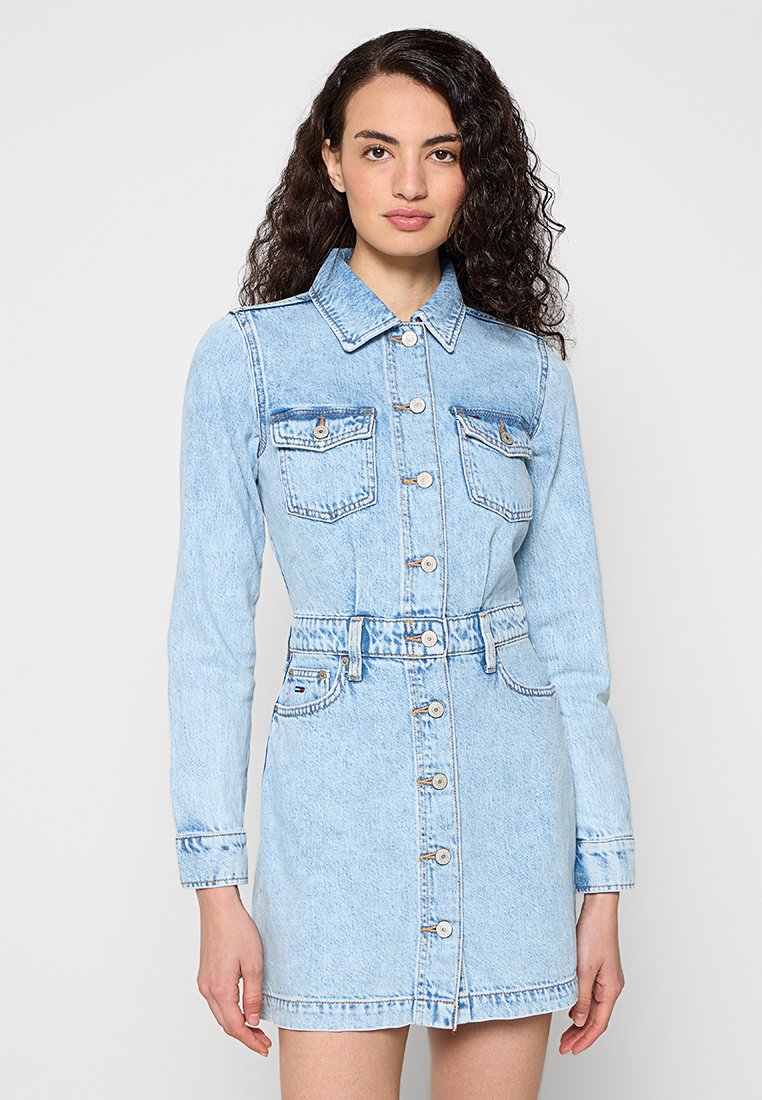 Frau mit lockigem Haar, die ein hellblaues Jeanskleid mit langen Ärmeln und Brusttaschen trägt und vor einem schlichten Hintergrund steht.