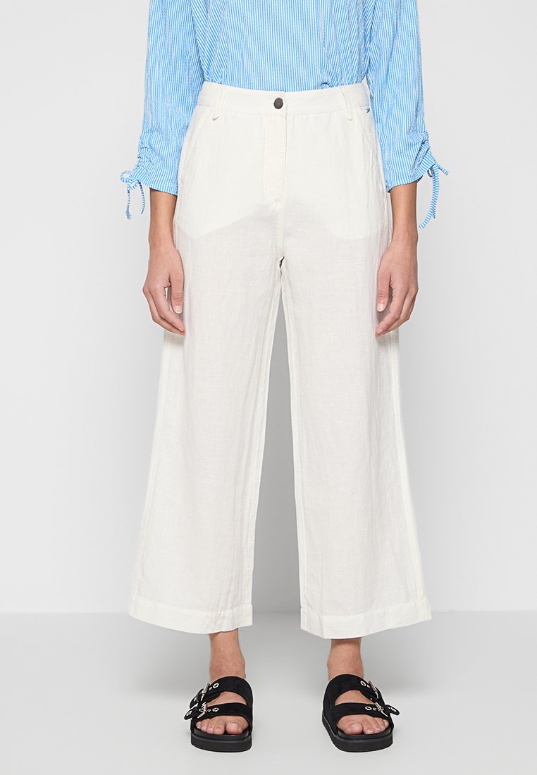 Weite, cremefarbene Leinenhosen mit hoher Taille, Ausstattung mit Knopfverschluss und Seitentaschen. Kombiniert mit schwarzen Slipper-Sandalen.