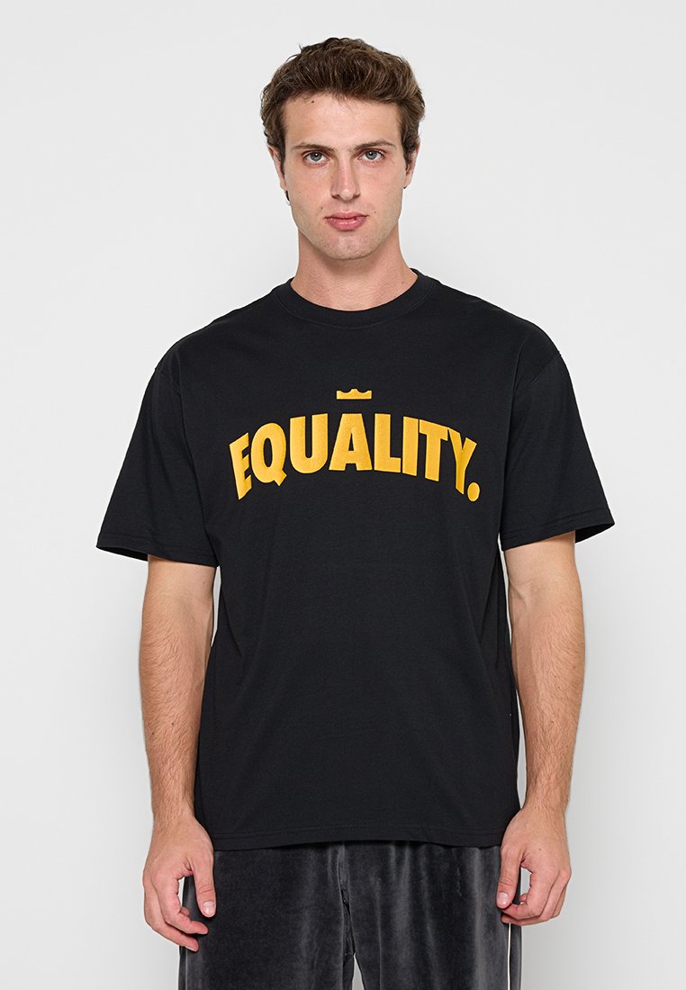 Junger Mann trägt ein schwarzes T-Shirt mit fettem gelbem Schriftzug „GLEICHHEIT“ und einem kleinen Kronensymbol über den Buchstaben, steht vor einem schlichten Hintergrund.