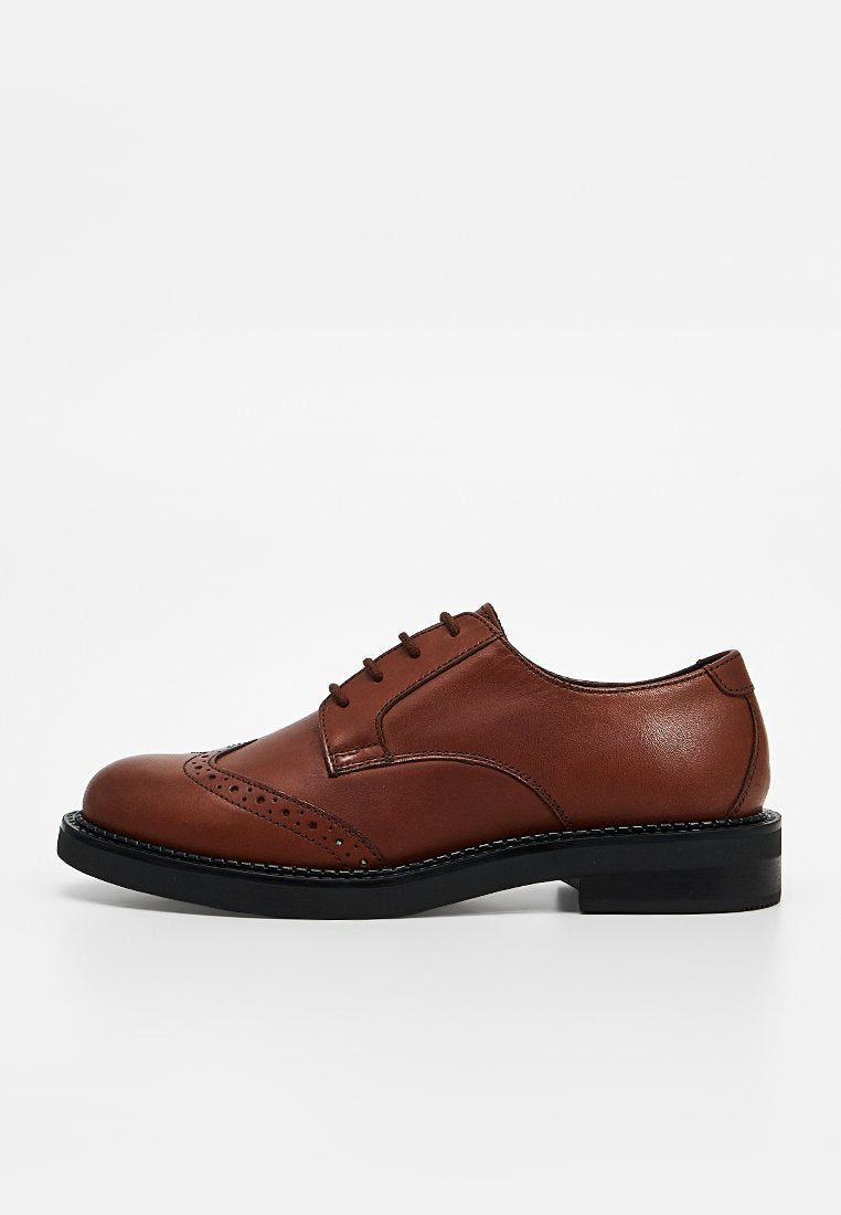 Brauner Lederschuh im Brogue-Stil mit schwarzer Sohle und Schnürsenkeln, Seitenansicht vor schlichtem hellem Hintergrund.